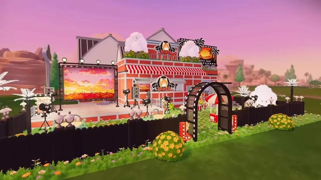 Wide shot of Drive‑In Cinema festival build with large screen, marquee building, film‑arch entrance, fencing and flower beds.